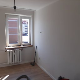 Met -Bud - Pomalowany na biało pokój z oknem, grzejnikiem i odkurzaczem w rogu, widok na budynek z zewnątrz.