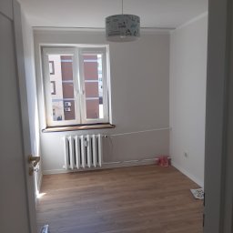 Met -Bud - Wnętrze pokoju po remoncie: świeżo pomalowane białe ściany, jasna drewniana podłoga, biały grzejnik pod oknem z widokiem na budynek, lampa z motywem dziecięcym. Widok zza otwartych drzwi.