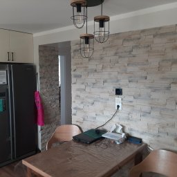 Met -Bud - Ściana w salonie wykończona dekoracyjną okładziną z kamienia imitującego cegłę, z widocznymi gniazdkami elektrycznymi i wiszącymi lampami w stylu industrialnym, stół z laptopem i krzesłami w tle...