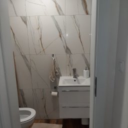 Met -Bud - Wykończona łazienka z białymi płytkami imitującymi marmur, białą szafką z umywalką, toaletą i drewnopodobną podłogą, widok zza framugi drzwi.