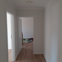 Met -Bud - Biały korytarz z drewnianą podłogą, widoczne otwarte drzwi i narzędzia malarskie w tle, oraz puszka elektryczna na podłodze przy ścianie.