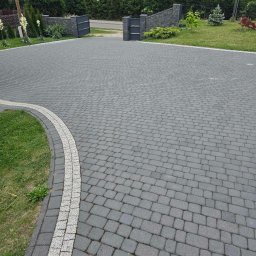 Met -Bud - Szeroki podjazd z szarej kostki brukowej ułożonej we wzór jodełki, obrzeża wykończone jasną kostką, brama wjazdowa w tle, zadbany trawnik po bokach.