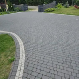 Szeroki podjazd z szarej kostki brukowej ułożonej we wzór jodełki, obrzeża wykończone jasną kostką, brama wjazdowa w tle, zadbany trawnik po bokach.