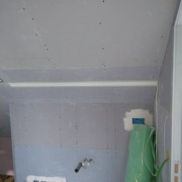 Met -Bud - Wnętrze w trakcie prac wykończeniowych: płyty gipsowo-kartonowe na ścianach i suficie, widoczne przewody elektryczne wystające ze ściany, rura kanalizacyjna oraz zielona folia ochronna przy ścianie.