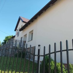 Met -Bud - Świeżo otynkowany, jasny dom z ciemnobrązowym dachem, widoczny zza metalowego ogrodzenia z ostrymi zakończeniami, fragment kamiennej podmurówki i balkonu.