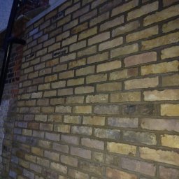 Met -Bud - Świeżo wymurowana ściana z cegły klinkierowej w odcieniach beżu i brązu, widoczna z boku, z ciemnymi spoinami i elementami rusztowania w tle.