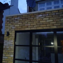 Met -Bud - Ściana z jasnej cegły klinkierowej z czarną ramą okienną, widoczny fragment elewacji domu z anteną satelitarną na dachu.