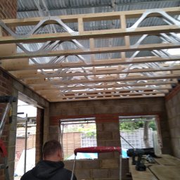 Met -Bud - Wnętrze w trakcie budowy domu murowanego z widoczną konstrukcją drewnianą stropu i metalowymi wzmocnieniami, ceglane ściany z oknami, drabiny i narzędzia budowlane.