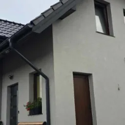 Fragment elewacji domu z szarą strukturą tynku, widoczne okno z kwiatami, drewniane drzwi wejściowe, ławka i czarne rynny.