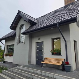 Met -Bud - Szary dom jednorodzinny z ciemną dachówką, drewnianą ławką przed wejściem i kwiatami w donicach, widok z zewnątrz na schody wejściowe wyłożone kostką brukową.