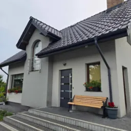 Szary dom jednorodzinny z ciemną dachówką, drewnianą ławką przed wejściem i kwiatami w donicach, widok z zewnątrz na schody wejściowe wyłożone kostką brukową.