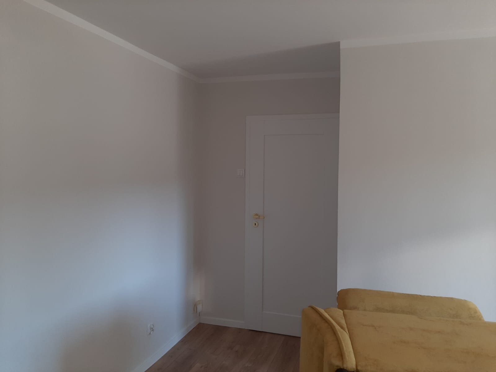 Jasne, świeżo pomalowane ściany w pokoju z białą listwą przysufitową i drzwiami. Fragment żółtej sofy w prawym dolnym rogu.