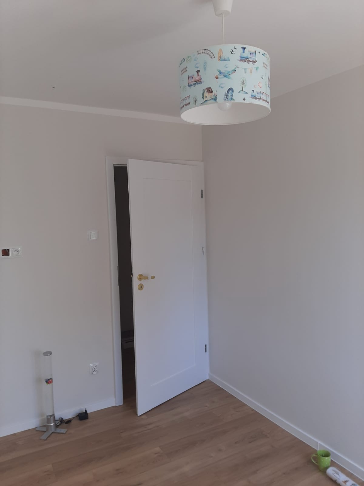 Wnętrze pokoju po odmalowaniu ścian na biało, widoczna biała ościeżnica z otwartymi drzwiami, drewniana podłoga oraz lampa sufitowa z motywem pociągów i samolotów.