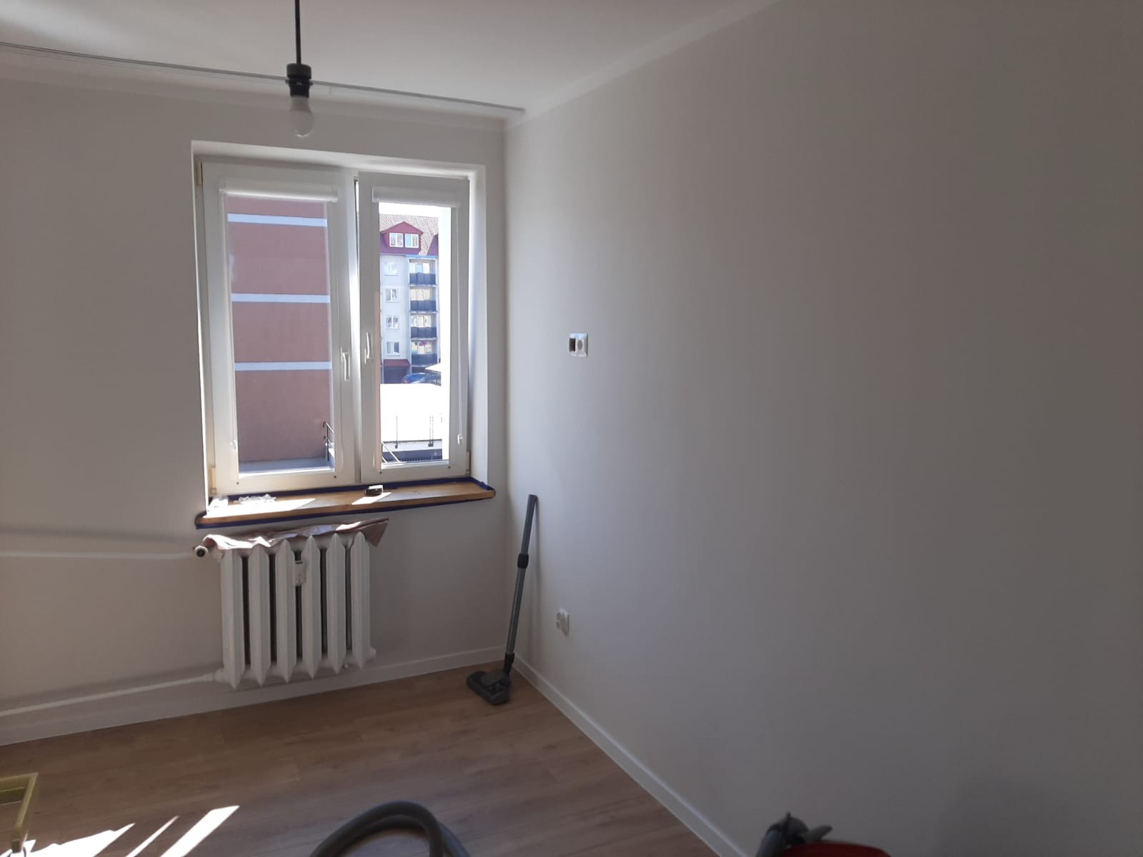 Pomalowany na biało pokój z oknem, grzejnikiem i odkurzaczem w rogu, widok na budynek z zewnątrz.