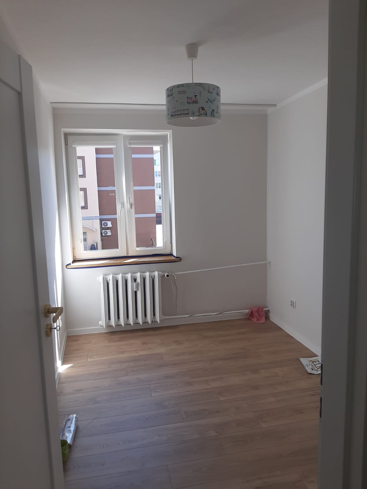 Wnętrze pokoju po remoncie: świeżo pomalowane białe ściany, jasna drewniana podłoga, biały grzejnik pod oknem z widokiem na budynek, lampa z motywem dziecięcym. Widok zza otwartych drzwi.