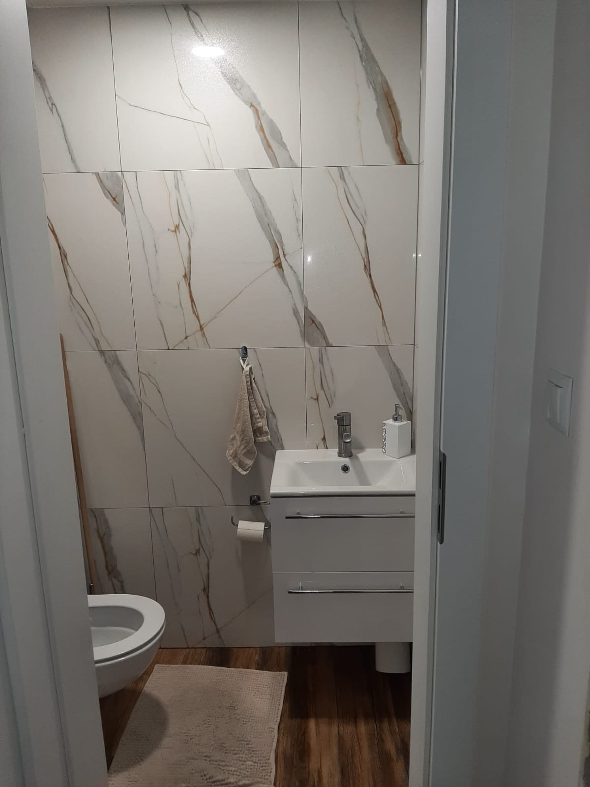 Wykończona łazienka z białymi płytkami imitującymi marmur, białą szafką z umywalką, toaletą i drewnopodobną podłogą, widok zza framugi drzwi.