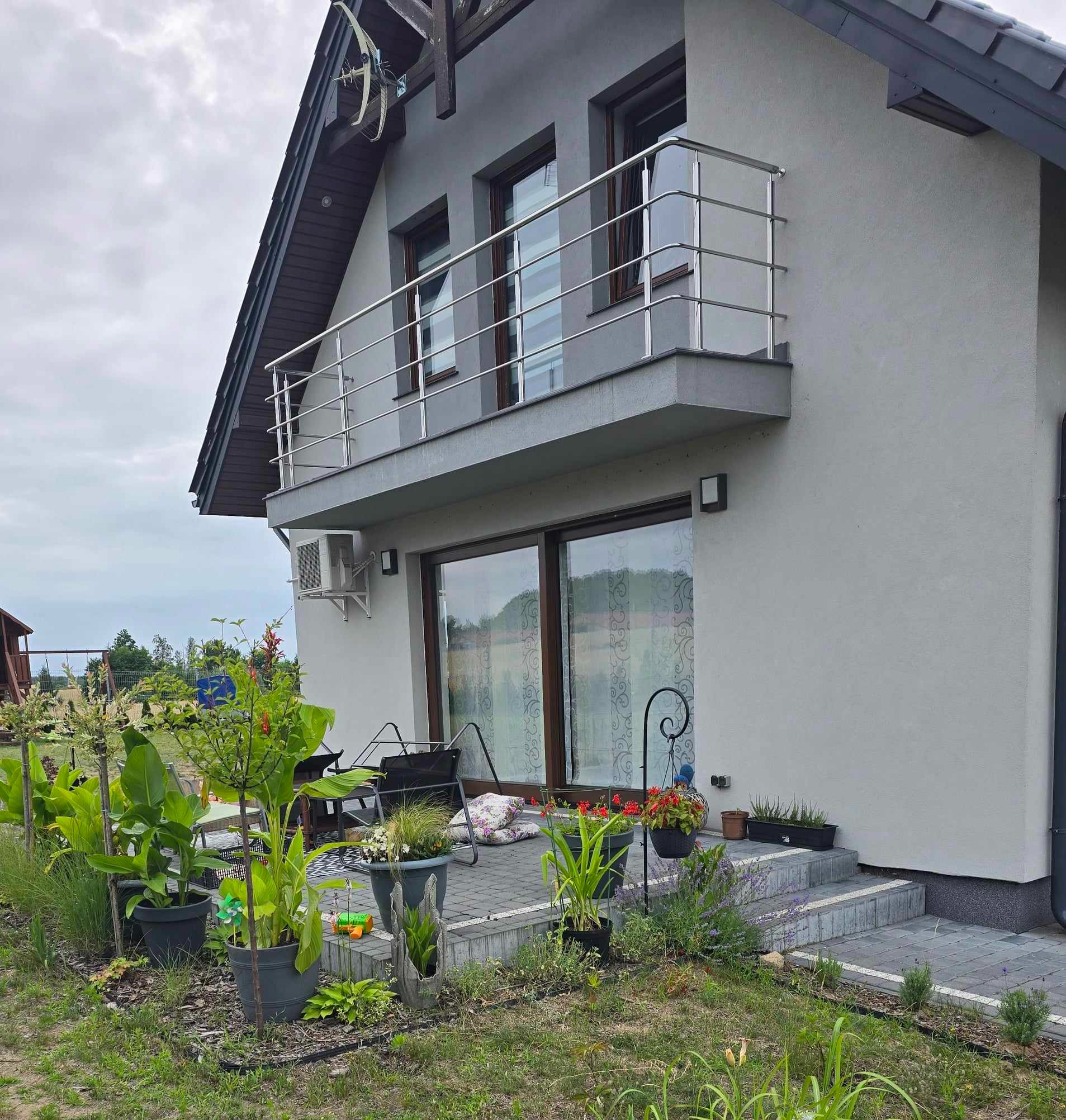 Szary dom jednorodzinny z balkonem i dużymi przesuwnymi drzwiami tarasowymi, widok z ogrodu z roślinami w donicach.