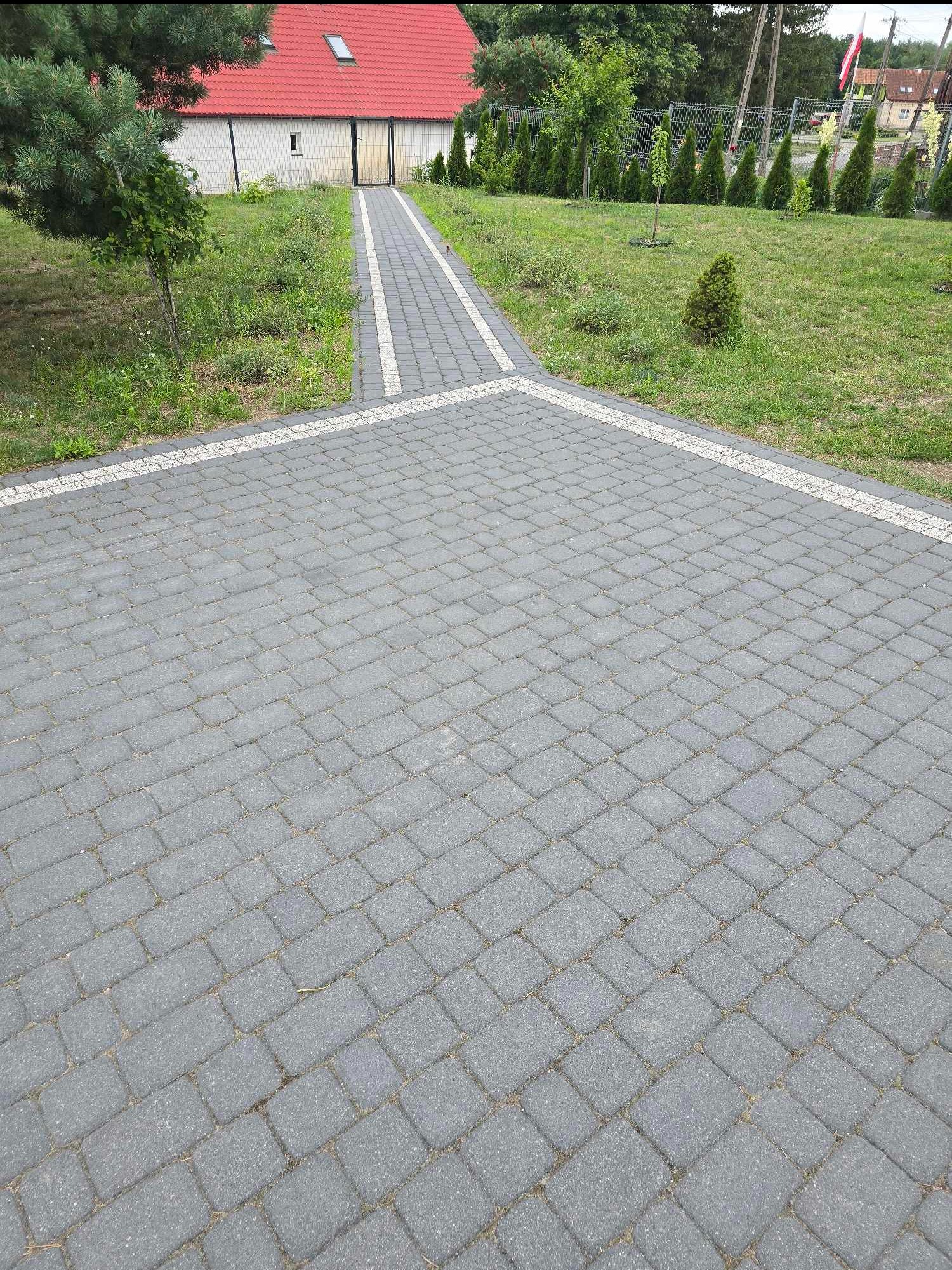 Ułożona szara kostka brukowa na podjeździe i ścieżce prowadzącej do budynku z czerwonym dachem, obrzeża z jasnej kostki, widoczne trawniki i drzewa.