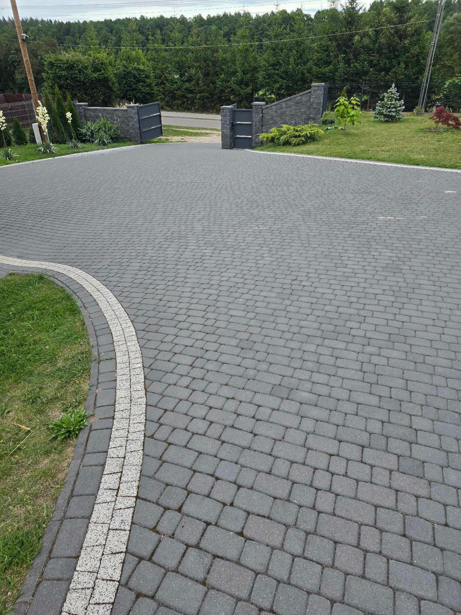 Szeroki podjazd z szarej kostki brukowej ułożonej we wzór jodełki, obrzeża wykończone jasną kostką, brama wjazdowa w tle, zadbany trawnik po bokach.