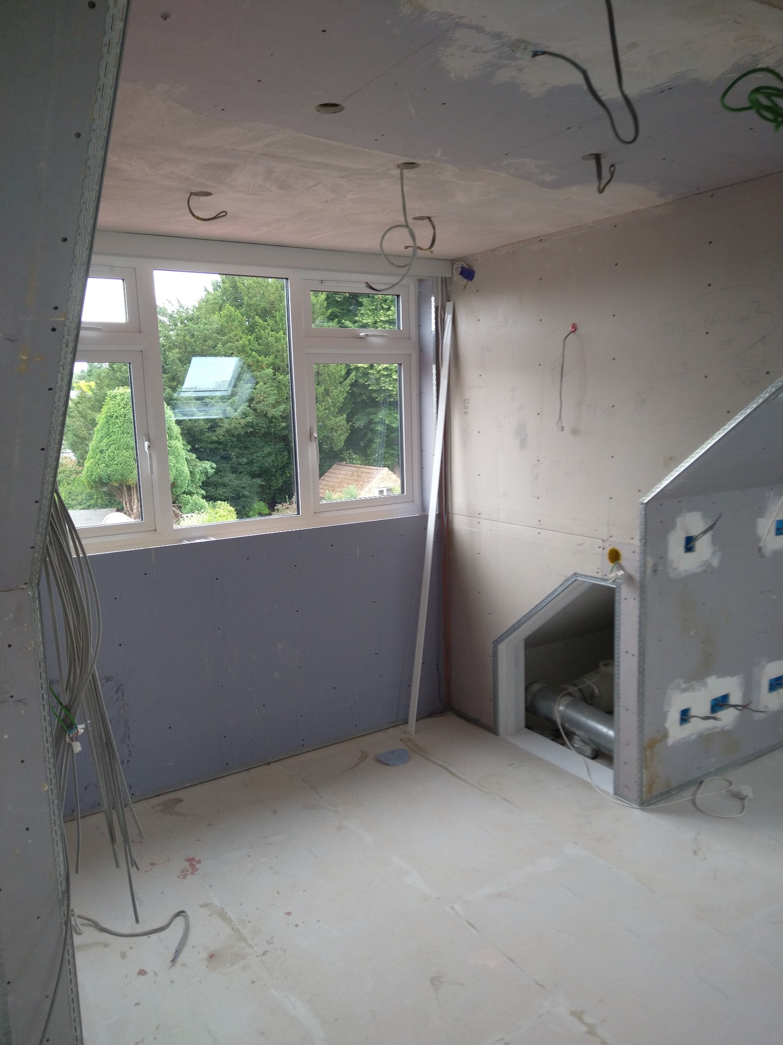 Wnętrze pokoju w trakcie wykańczania: widoczne płyty gipsowo-kartonowe na ścianach i suficie, okno z widokiem na zieleń, wystające przewody elektryczne i fragment instalacji wentylacyjnej w zabudowie.