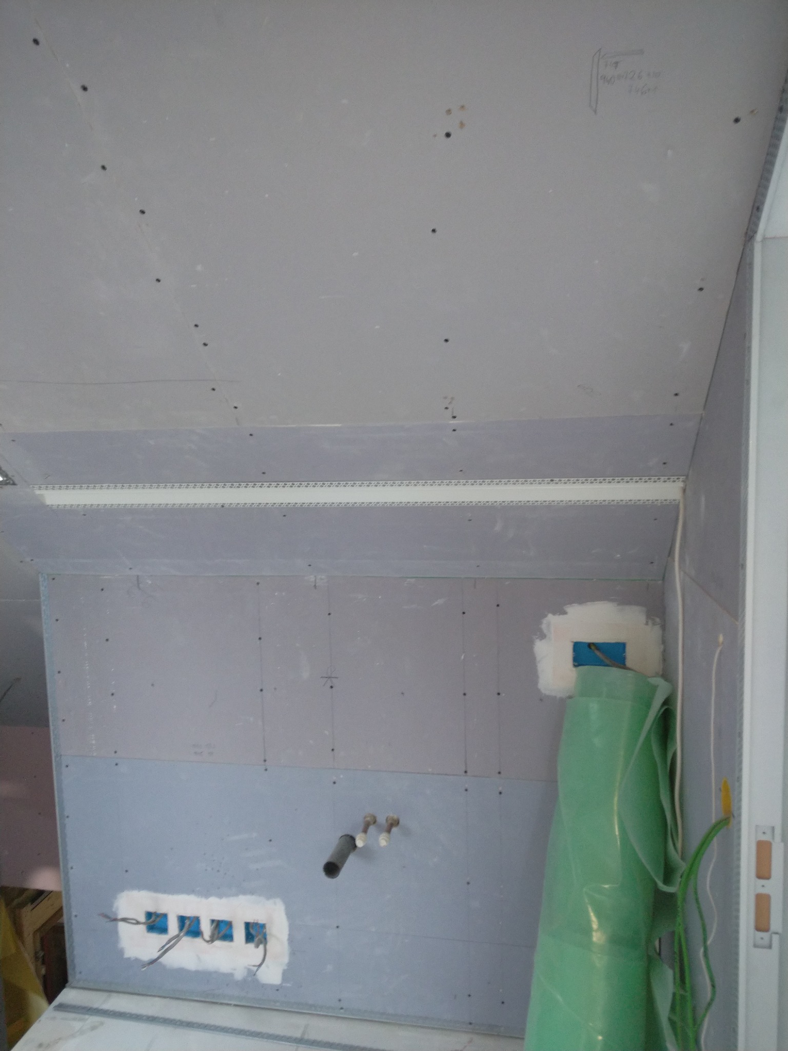 Wnętrze w trakcie prac wykończeniowych: płyty gipsowo-kartonowe na ścianach i suficie, widoczne przewody elektryczne wystające ze ściany, rura kanalizacyjna oraz zielona folia ochronna przy ścianie.