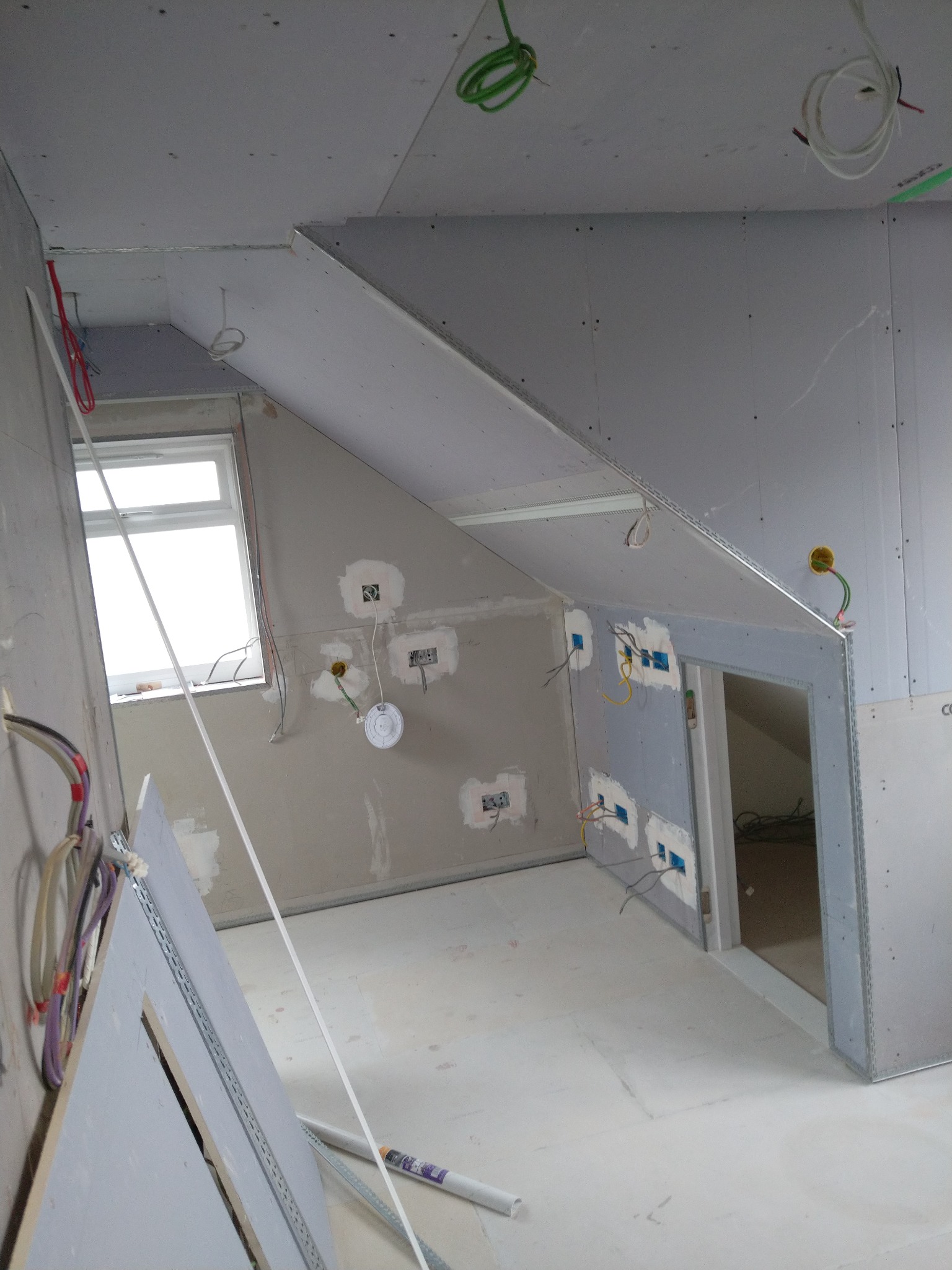 Wnętrze poddasza w trakcie prac wykończeniowych: zamontowane płyty gipsowo-kartonowe na ścianach i skosach, widoczne instalacje elektryczne, przygotowane otwory na gniazdka, surowe wykończenie...