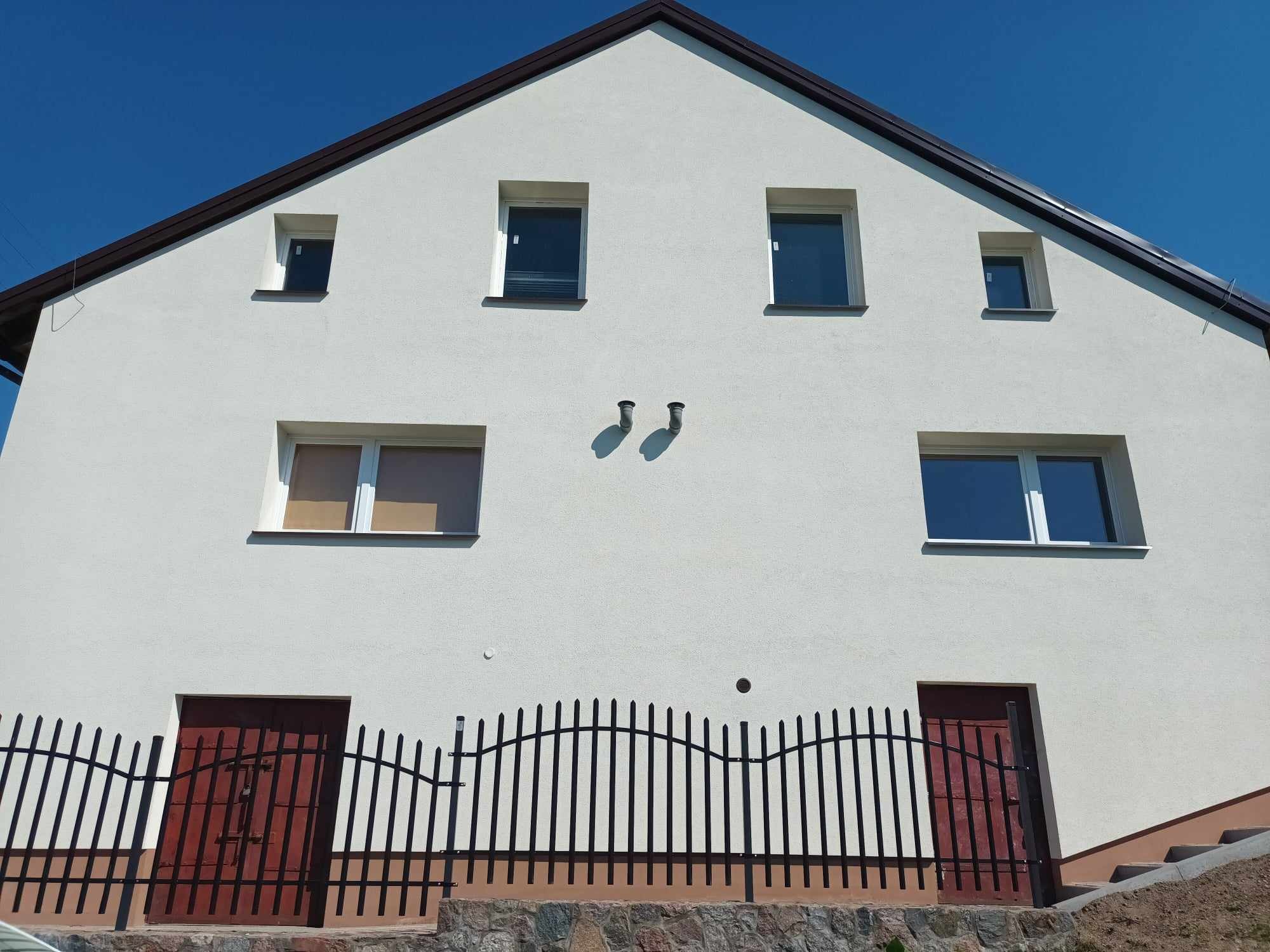 Dwukondygnacyjny dom z białą elewacją, brązowym dachem i czarnym ogrodzeniem, widoczny z dołu w słoneczny dzień. Dwie brązowe drzwi wejściowe na poziomie gruntu, ogrodzone czarnym metalowym płotem...