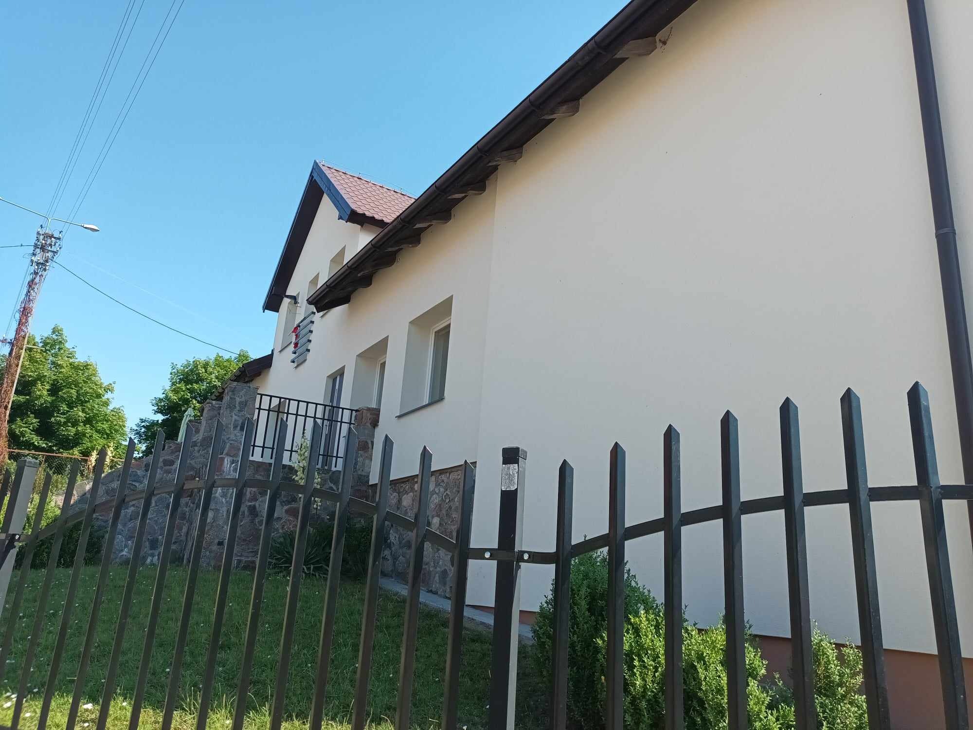 Świeżo otynkowany, jasny dom z ciemnobrązowym dachem, widoczny zza metalowego ogrodzenia z ostrymi zakończeniami, fragment kamiennej podmurówki i balkonu.