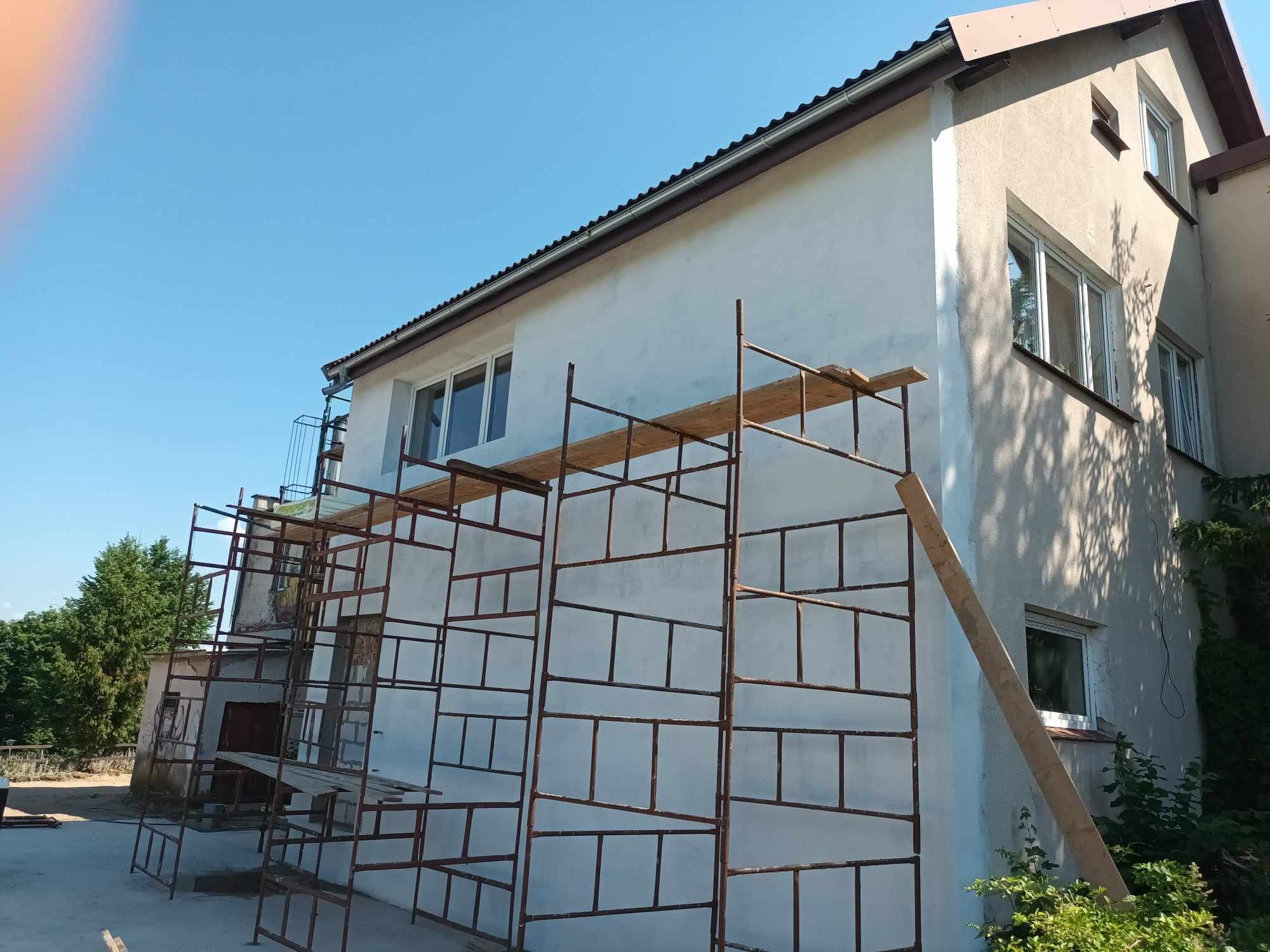 Budynek jednorodzinny z białymi ścianami w trakcie ocieplania, widoczne rusztowania i deski, fragment elewacji jeszcze nieocieplony.