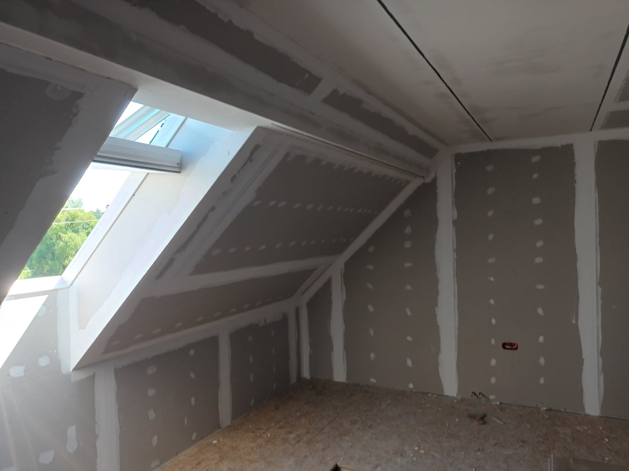 Poddasze w trakcie wykańczania płytami gipsowo-kartonowymi, widoczne łączenia szpachlowane, okno dachowe i surowe podłoże podłogi.