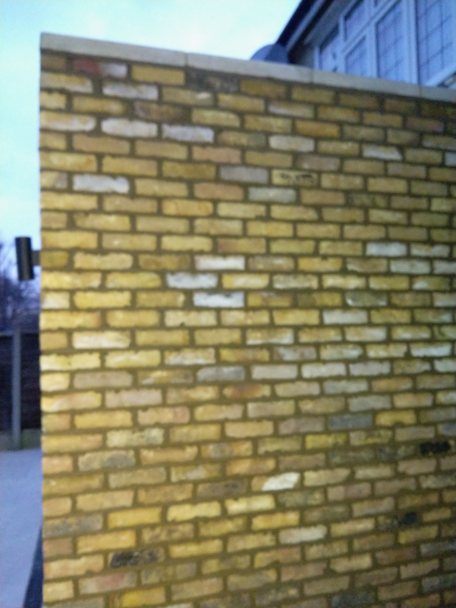 Świeżo wymurowana ściana z żółtej cegły, widoczny fragment budynku w tle, pochmurne niebo.