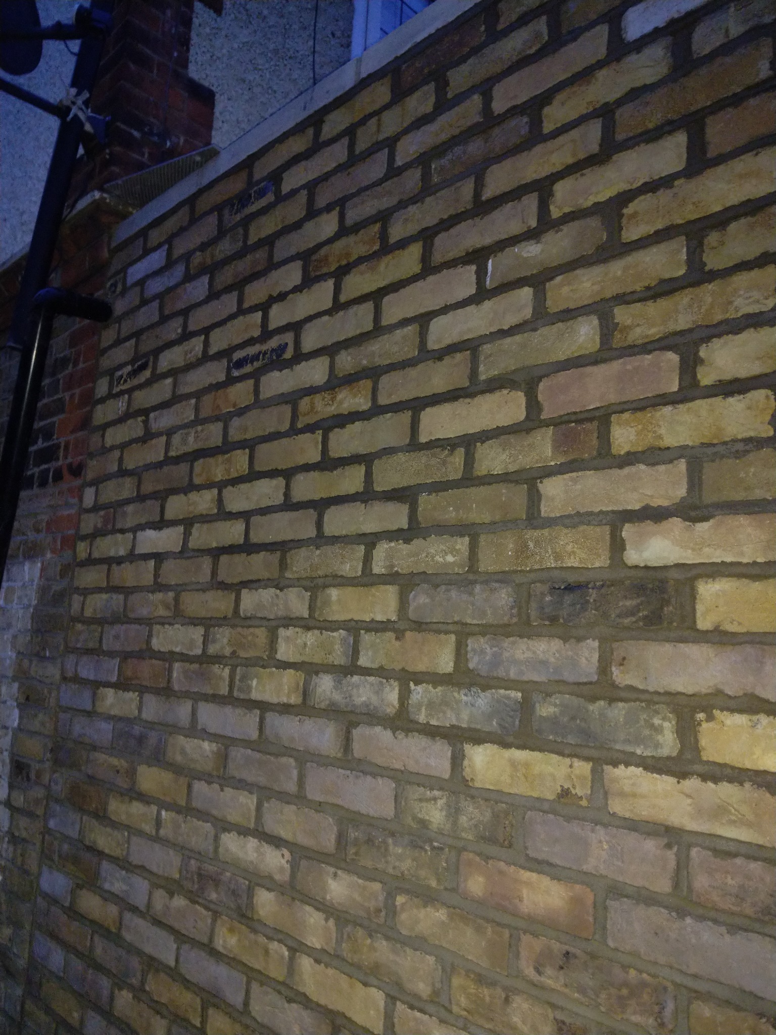 Świeżo wymurowana ściana z cegły klinkierowej w odcieniach beżu i brązu, widoczna z boku, z ciemnymi spoinami i elementami rusztowania w tle.