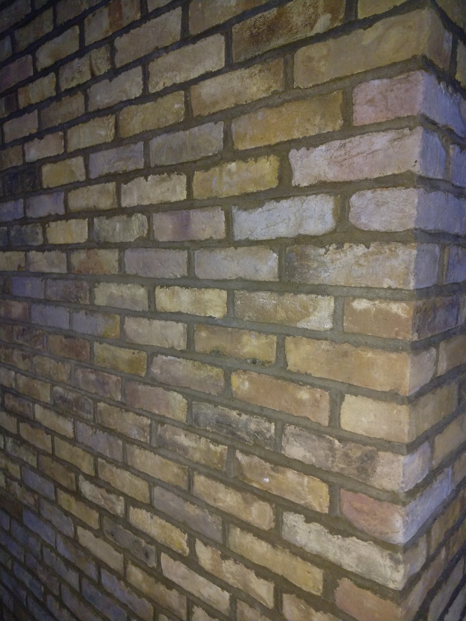 Fragment narożnej ściany z cegły klinkierowej w odcieniach beżu, brązu i szarości, z widocznymi spoinami, oświetlonej z dwóch stron, tworzącej efekt cienia na jednej z płaszczyzn.