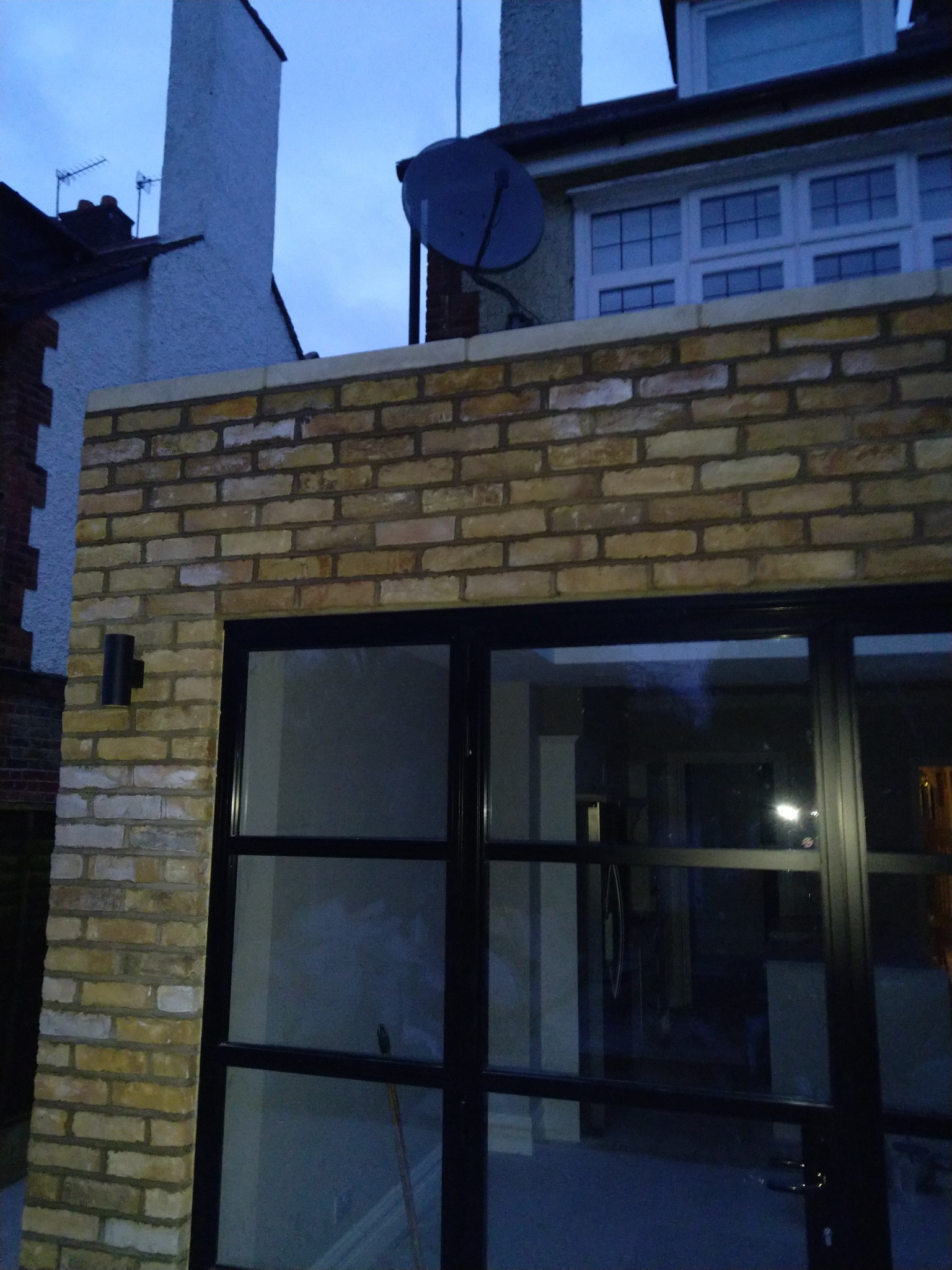 Ściana z jasnej cegły klinkierowej z czarną ramą okienną, widoczny fragment elewacji domu z anteną satelitarną na dachu.