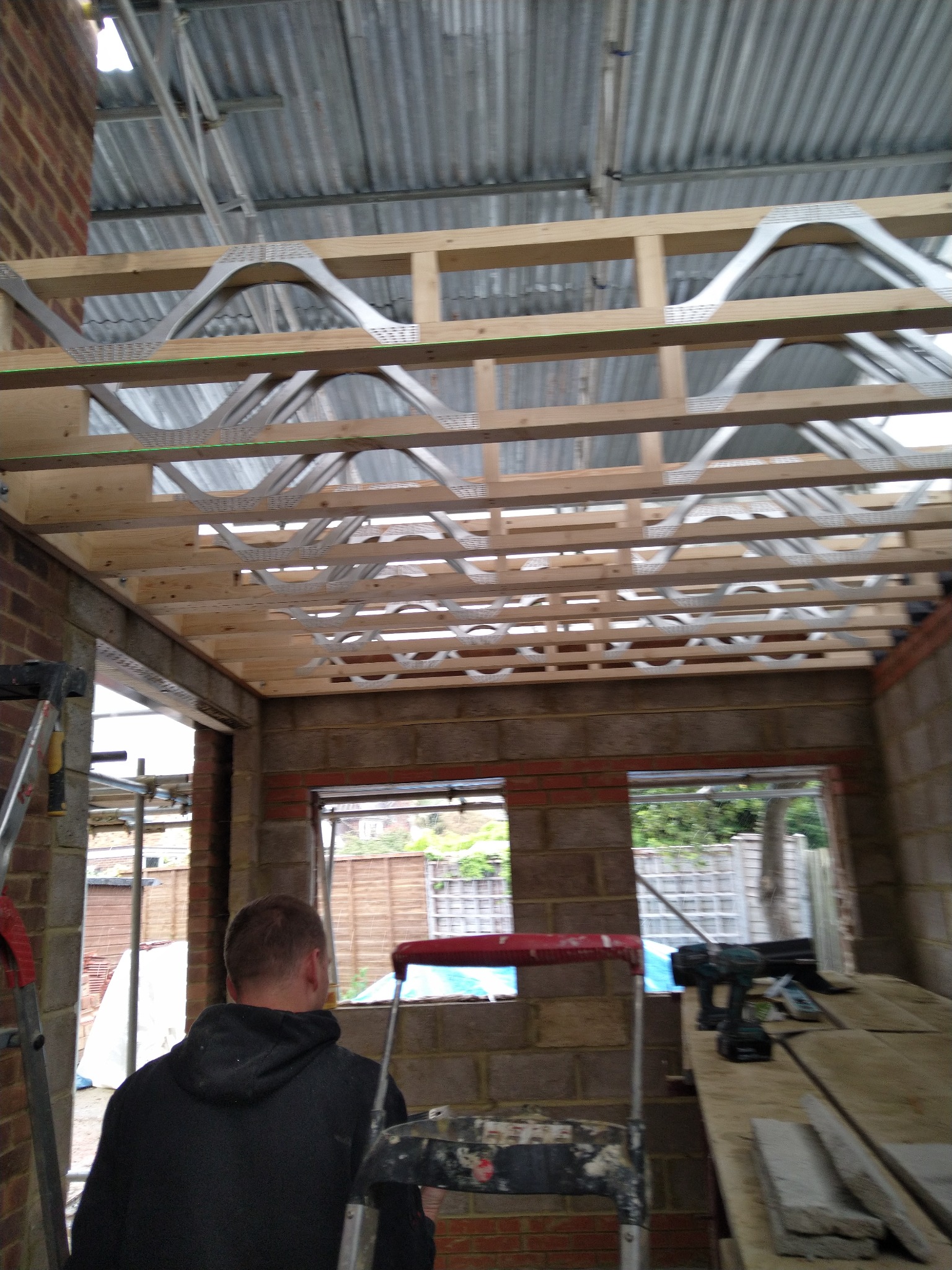 Wnętrze w trakcie budowy domu murowanego z widoczną konstrukcją drewnianą stropu i metalowymi wzmocnieniami, ceglane ściany z oknami, drabiny i narzędzia budowlane.