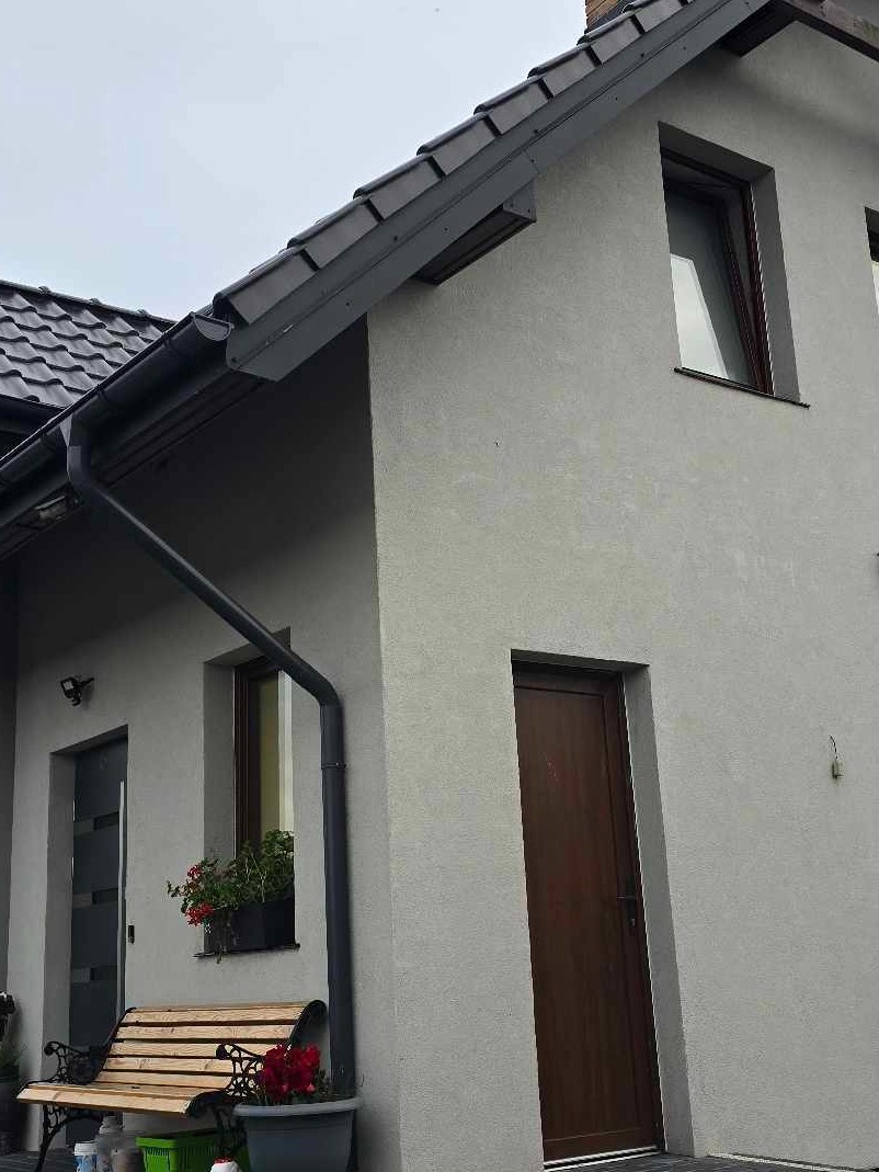 Fragment elewacji domu z szarą strukturą tynku, widoczne okno z kwiatami, drewniane drzwi wejściowe, ławka i czarne rynny.