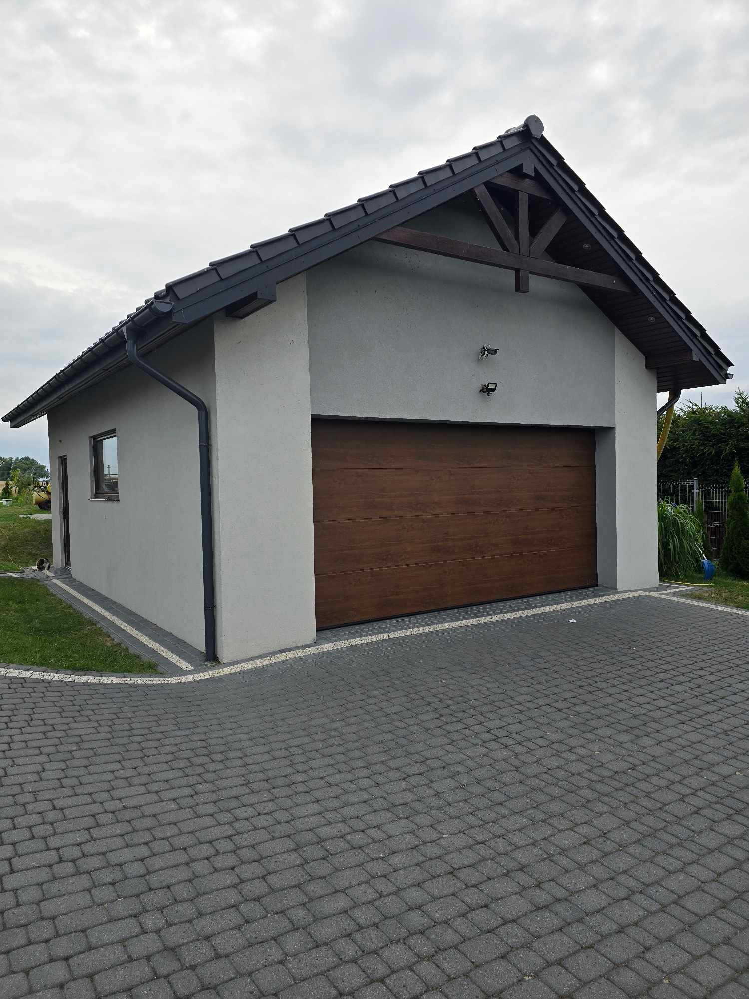 Nowoczesny, parterowy garaż z brązową bramą segmentową, szarą elewacją i ciemną dachówką, wykończony kostką brukową.
