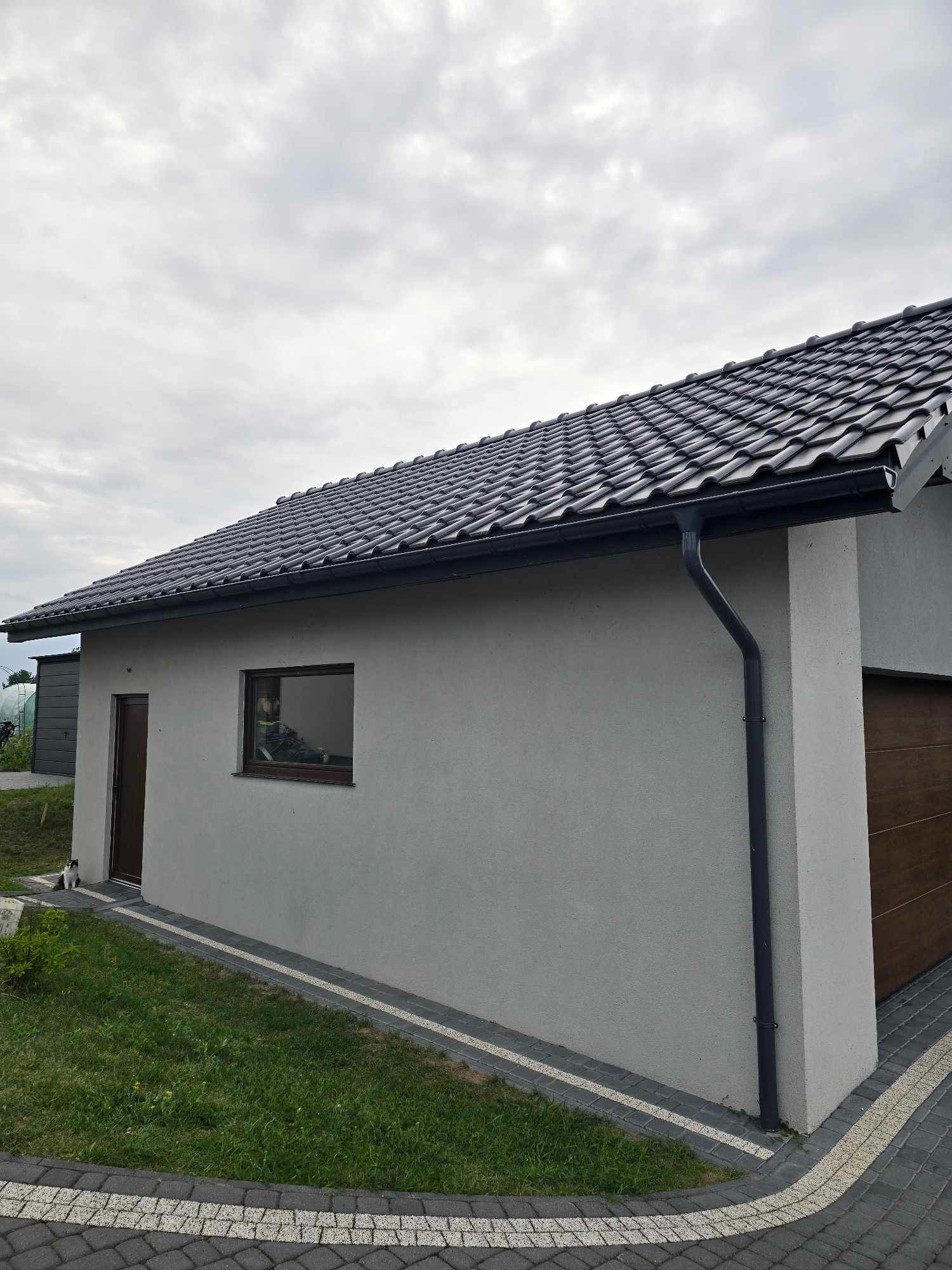 Nowoczesny dom jednorodzinny z szarą elewacją, grafitową dachówką i brązową bramą garażową, widoczne orynnowanie w kolorze grafitowym, na pierwszym planie trawnik i kostka brukowa, w oknie odbija...