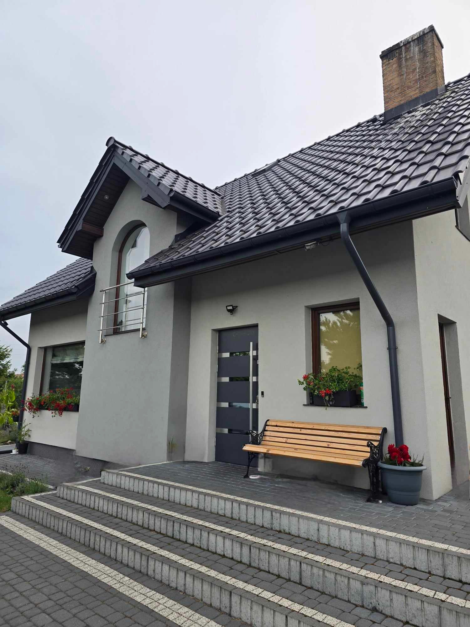 Szary dom jednorodzinny z ciemną dachówką, drewnianą ławką przed wejściem i kwiatami w donicach, widok z zewnątrz na schody wejściowe wyłożone kostką brukową.