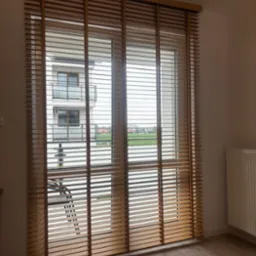 Zamknięte poziome lamele w oknie z widokiem na balkon sąsiedniego budynku i krajobraz za nim, ciepłe odcienie drewna i wyraźna faktura.