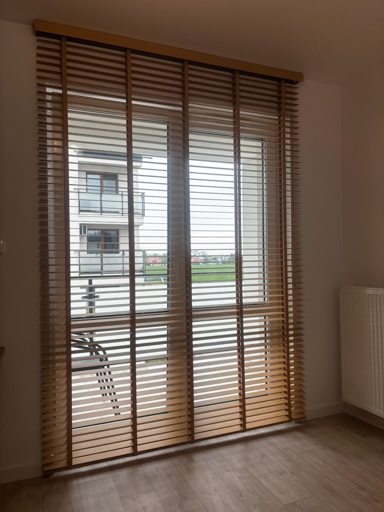 Zamknięte poziome lamele w oknie z widokiem na balkon sąsiedniego budynku i krajobraz za nim, ciepłe odcienie drewna i wyraźna faktura.