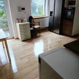 Błyszcząca drewnopodobna podłoga w kuchni z widocznym fragmentem stołu, szafki z szufladami, krzesła i nowoczesnej zabudowy kuchennej ze sprzętem AGD.