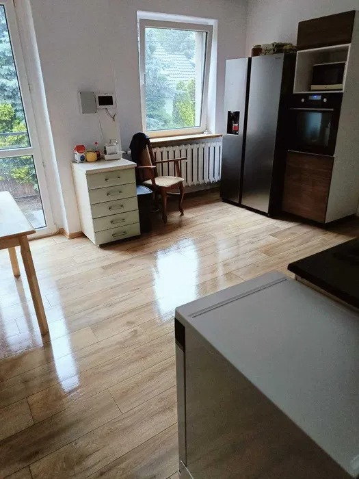 Błyszcząca drewnopodobna podłoga w kuchni z widocznym fragmentem stołu, szafki z szufladami, krzesła i nowoczesnej zabudowy kuchennej ze sprzętem AGD.