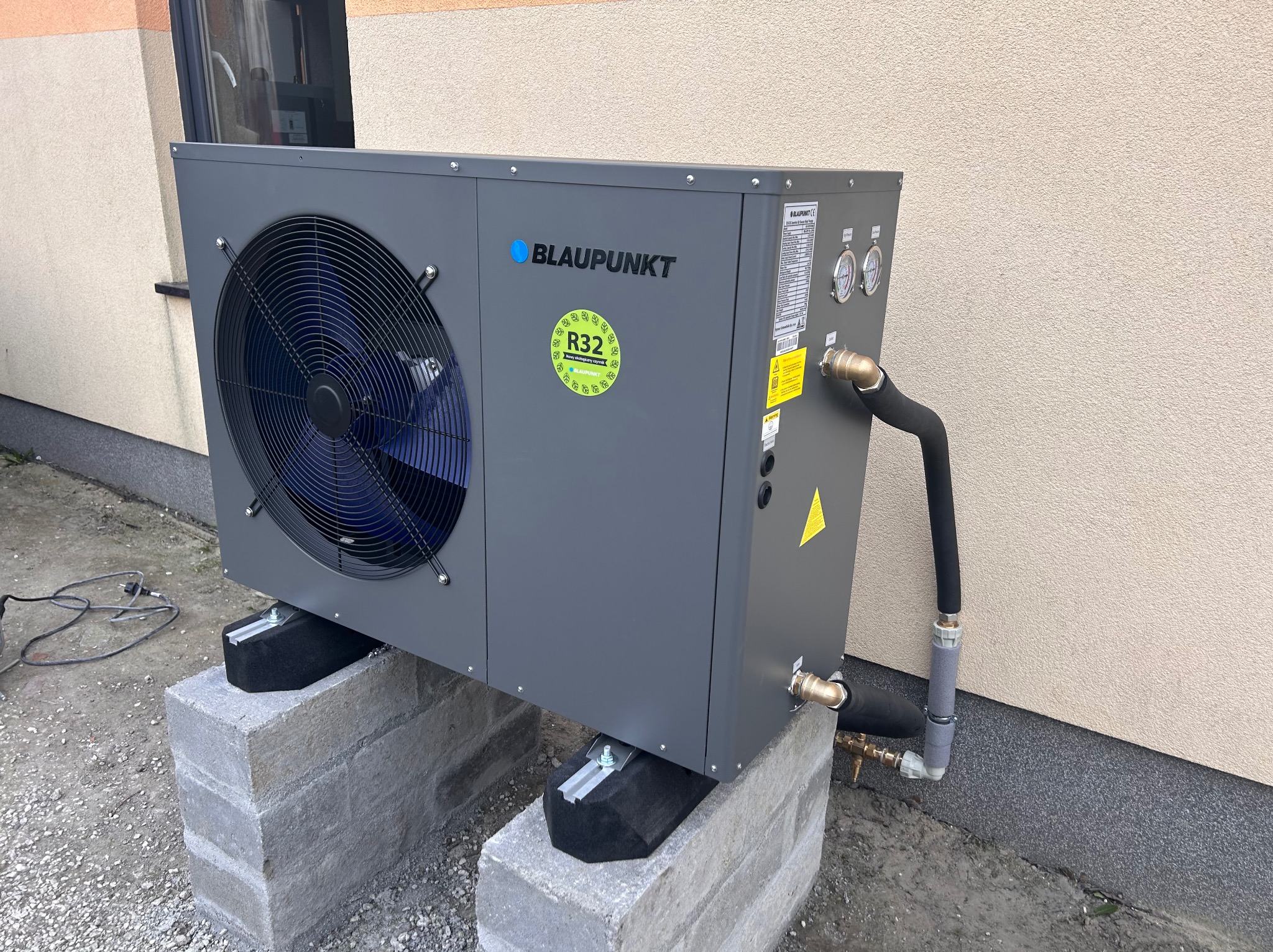 Szara pompa ciepła marki Blaupunkt, model z czynnikiem chłodniczym R32, umieszczona na betonowych bloczkach z czarnymi podkładkami tłumiącymi drgania, zamontowana przy elewacji budynku.