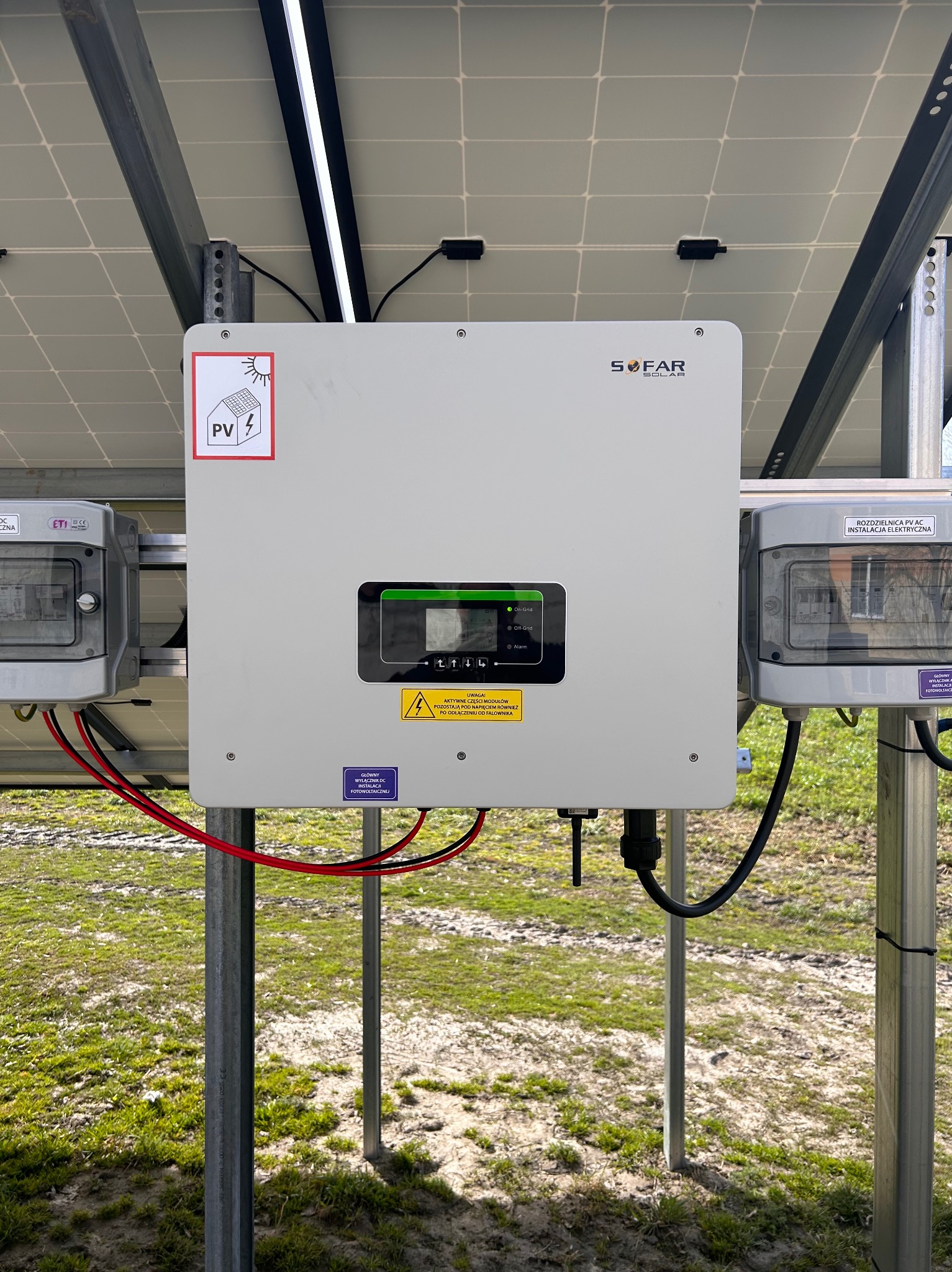 Szary inwerter SOFAR SOLAR zamontowany na metalowych słupkach, pod panelami słonecznymi, z widoczną tabliczką ostrzegawczą o napięciu i rozdzielnicą PV AC z boku.