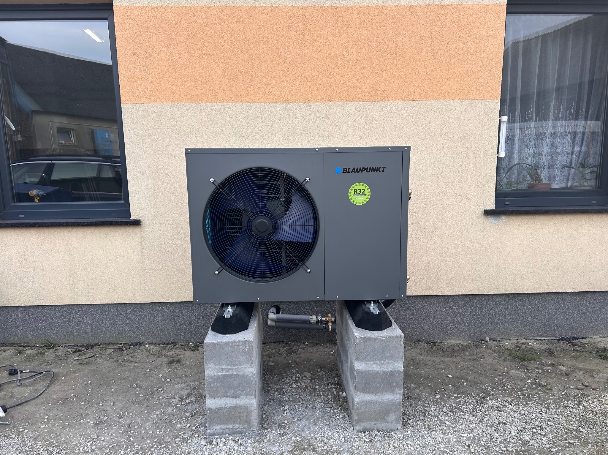 Szara jednostka zewnętrzna pompy ciepła marki Blaupunkt, zamontowana na betonowych bloczkach przy ścianie budynku o dwukolorowej elewacji, z widocznymi oknami i fragmentem ogrodu.