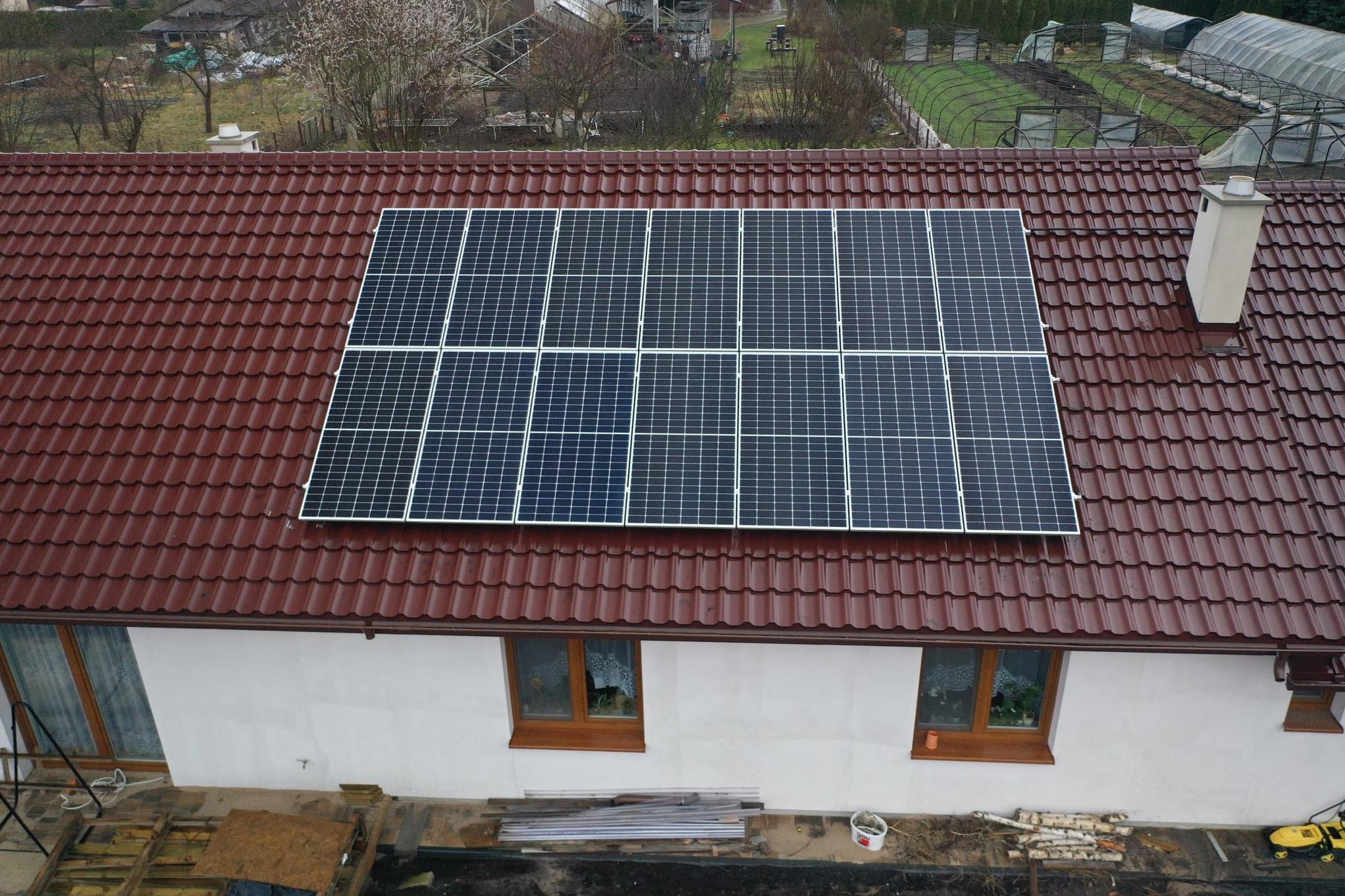 Panel słoneczny na dachu domu z brązową dachówką, widok z góry, w tle zabudowa jednorodzinna i szklarnie.