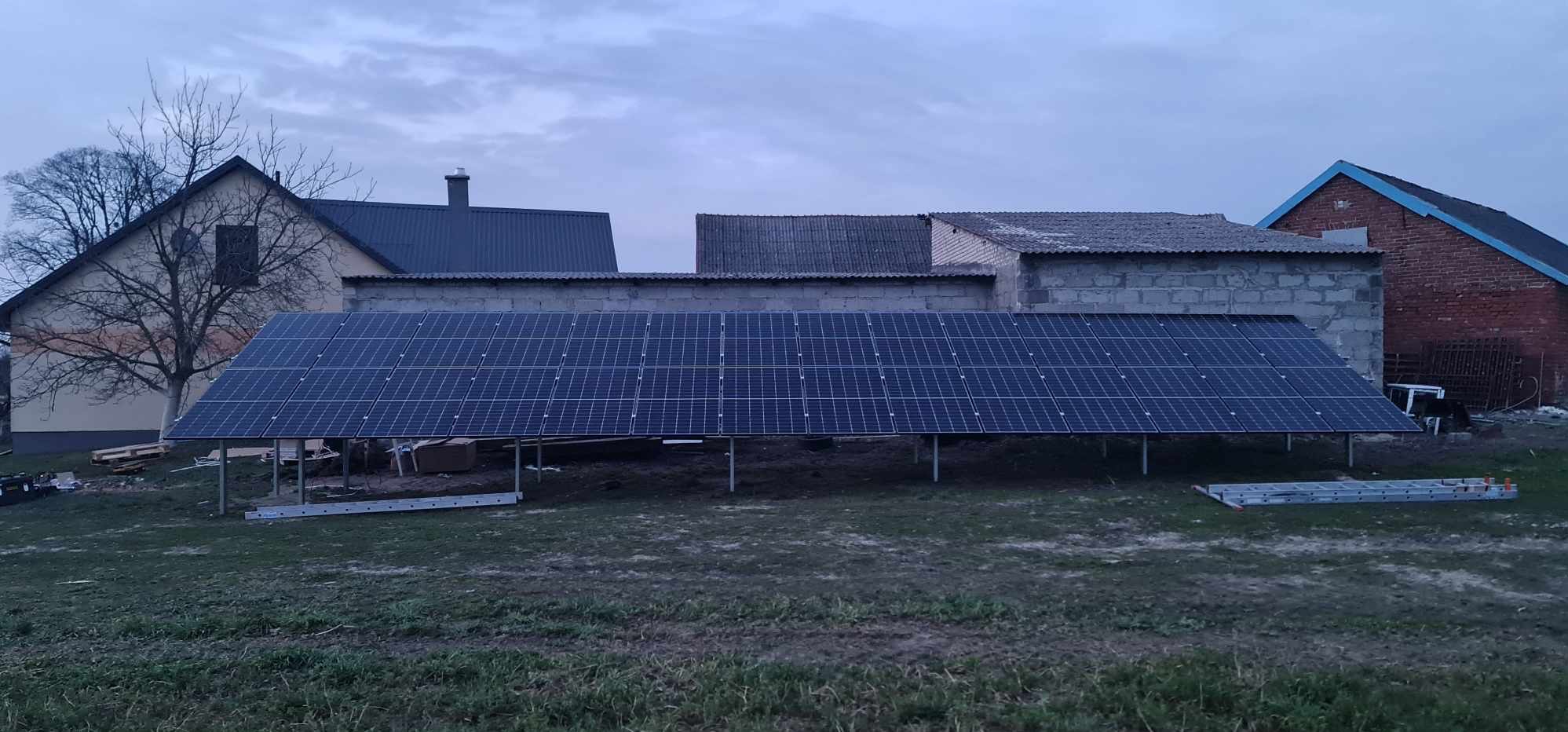 Instalacja paneli słonecznych na metalowej konstrukcji na tle budynków gospodarczych i domu jednorodzinnego, drabina leżąca na ziemi, pochmurne niebo.