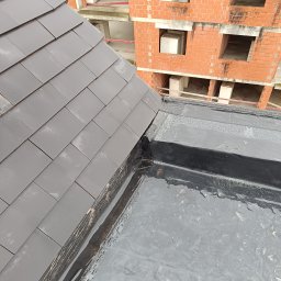 Usługi dekarskie Kamil Kowalski - Szare dachówki ceramiczne na nowym dachu, widoczne detale łączenia z czarną membraną hydroizolacyjną i budynek w trakcie budowy w tle.
