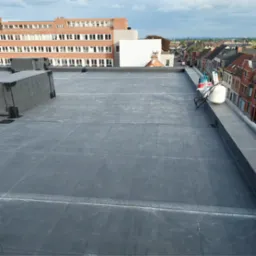 Wykończony, płaski dach pokryty czarną membraną EPDM, widoczne zgrzewy i narzędzia dekarskie na krawędzi dachu, budynki w tle.